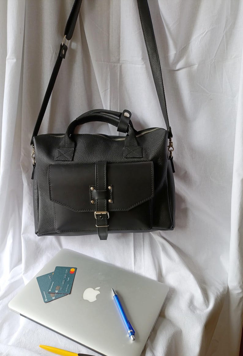 Puede incluir: Un bolso tipo satchel de cuero negro con solapa delantera y cierre de hebilla, asa superior y correa para el hombro desmontable. Tambi&eacute;n se ven un port&aacute;til, tarjetas de cr&eacute;dito y un bol&iacute;grafo.