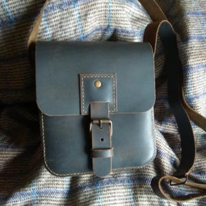 Pu&ograve; includere: Una borsa a tracolla in pelle blu scuro con chiusura a fibbia e una tracolla marrone. La borsa &egrave; appoggiata su un tessuto a quadri.
