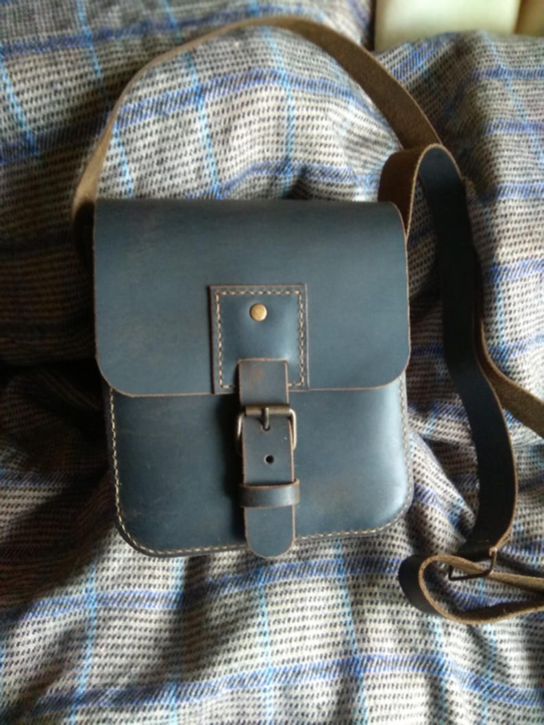 Pu&ograve; includere: Una borsa a tracolla in pelle blu scuro con chiusura a fibbia e una tracolla marrone. La borsa &egrave; appoggiata su un tessuto a quadri.