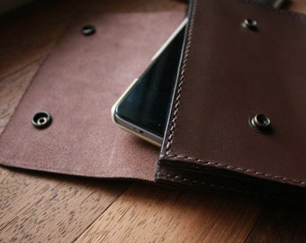 May include: A brown leather phone case with a flap that opens to reveal a phone inside. The case has stitching around the edges and metal snaps.