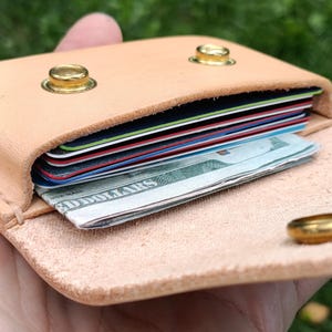 May include: A light tan leather card holder featuring two gold-toned metal snap buttons on the front flap. The interior compartment is filled with a stack of multi-colored credit cards and a folded United States dollar bill visible at the front.