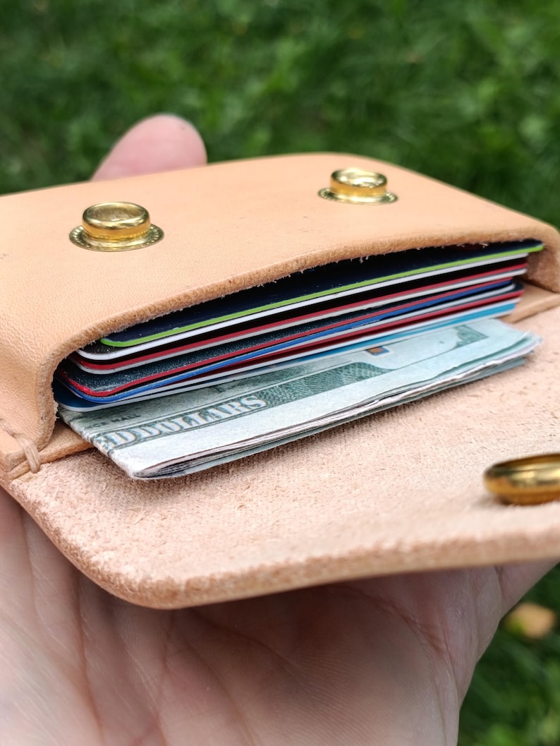May include: A light tan leather card holder featuring two gold-toned metal snap buttons on the front flap. The interior compartment is filled with a stack of multi-colored credit cards and a folded United States dollar bill visible at the front.