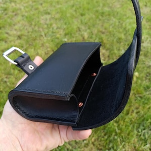 May include: Black leather case with a curved flap and a strap. The case is open, revealing a dark interior and copper-colored rivets. A silver-colored buckle is attached to the strap. The case is held against a background of green grass.