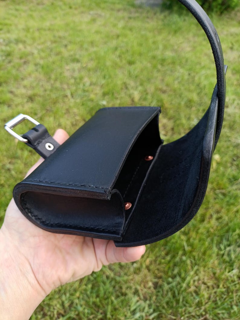 May include: Black leather case with a curved flap and a strap. The case is open, revealing a dark interior and copper-colored rivets. A silver-colored buckle is attached to the strap. The case is held against a background of green grass.