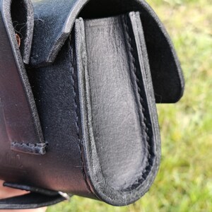 May include: Close-up of a black leather pouch with a curved flap. The pouch is stitched with black thread and has a loop on the side. The background is blurred green grass.