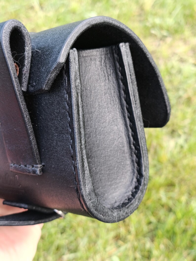 May include: Close-up of a black leather pouch with a curved flap. The pouch is stitched with black thread and has a loop on the side. The background is blurred green grass.