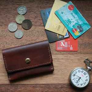 May include: A brown leather wallet with a gold snap, coins, credit cards, and a silver pocket watch with a chain are arranged on a wooden surface. The watch shows the time.