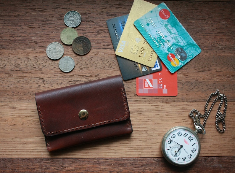 May include: A brown leather wallet with a gold snap, coins, credit cards, and a silver pocket watch with a chain are arranged on a wooden surface. The watch shows the time.