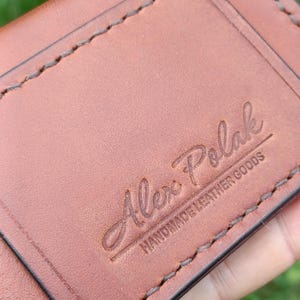 Puede incluir: Una cartera de cuero color canela con costuras marrones visibles a lo largo de los bordes. La superficie est&aacute; grabada con el texto Alex Polak y las palabras HANDMADE LEATHER GOODS debajo en una fuente sans-serif.