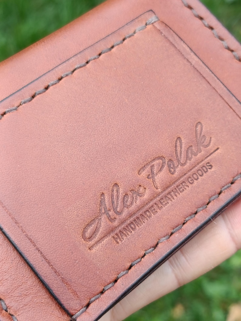 Puede incluir: Una cartera de cuero color canela con costuras marrones visibles a lo largo de los bordes. La superficie est&aacute; grabada con el texto Alex Polak y las palabras HANDMADE LEATHER GOODS debajo en una fuente sans-serif.