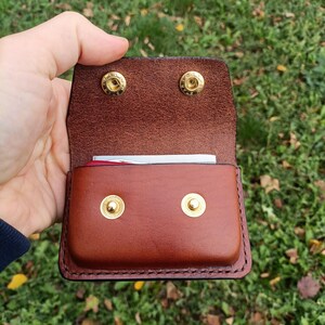May include: A brown leather pouch with a flap closure, featuring two gold-colored metal snap buttons on the front and two matching snap buttons on the interior flap. The leather has visible stitching along the edges and contains a small item.