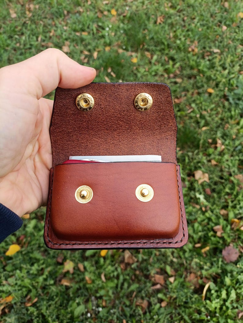 May include: A brown leather pouch with a flap closure, featuring two gold-colored metal snap buttons on the front and two matching snap buttons on the interior flap. The leather has visible stitching along the edges and contains a small item.