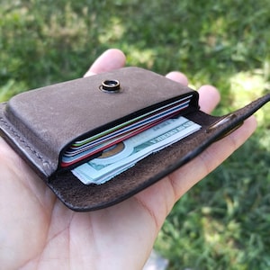 May include: A dark brown leather card holder featuring a single metal snap button closure. The wallet contains several stacked credit cards and a folded United States one hundred dollar bill visible within the main compartment slot.