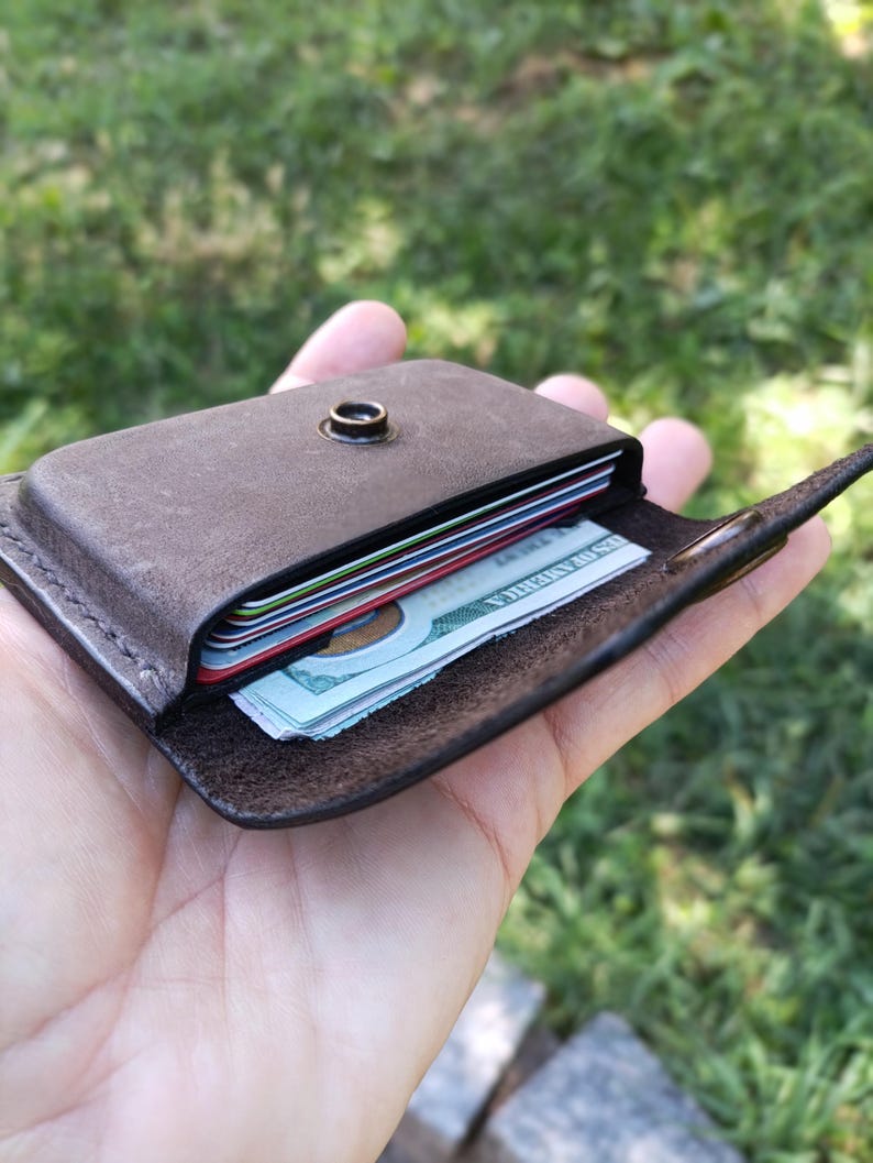 May include: A dark brown leather card holder featuring a single metal snap button closure. The wallet contains several stacked credit cards and a folded United States one hundred dollar bill visible within the main compartment slot.