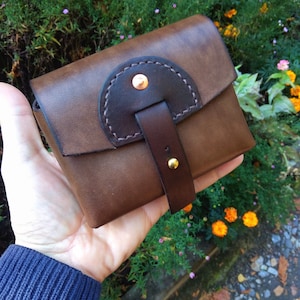 May include: A small brown leather pouch with a flap closure and a decorative strap. The pouch has a dark brown leather accent and a copper rivet.