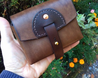May include: A small brown leather pouch with a flap closure and a decorative strap. The pouch has a dark brown leather accent and a copper rivet.
