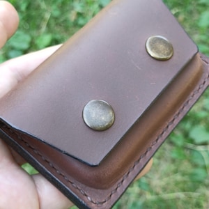 Puede incluir: Un tarjetero rectangular de cuero marr&oacute;n con cierre de solapa asegurado por dos broches met&aacute;licos redondos de color lat&oacute;n. Los bordes del cuero est&aacute;n cosidos con hilo oscuro y la superficie tiene un acabado mate suave con grano visible.