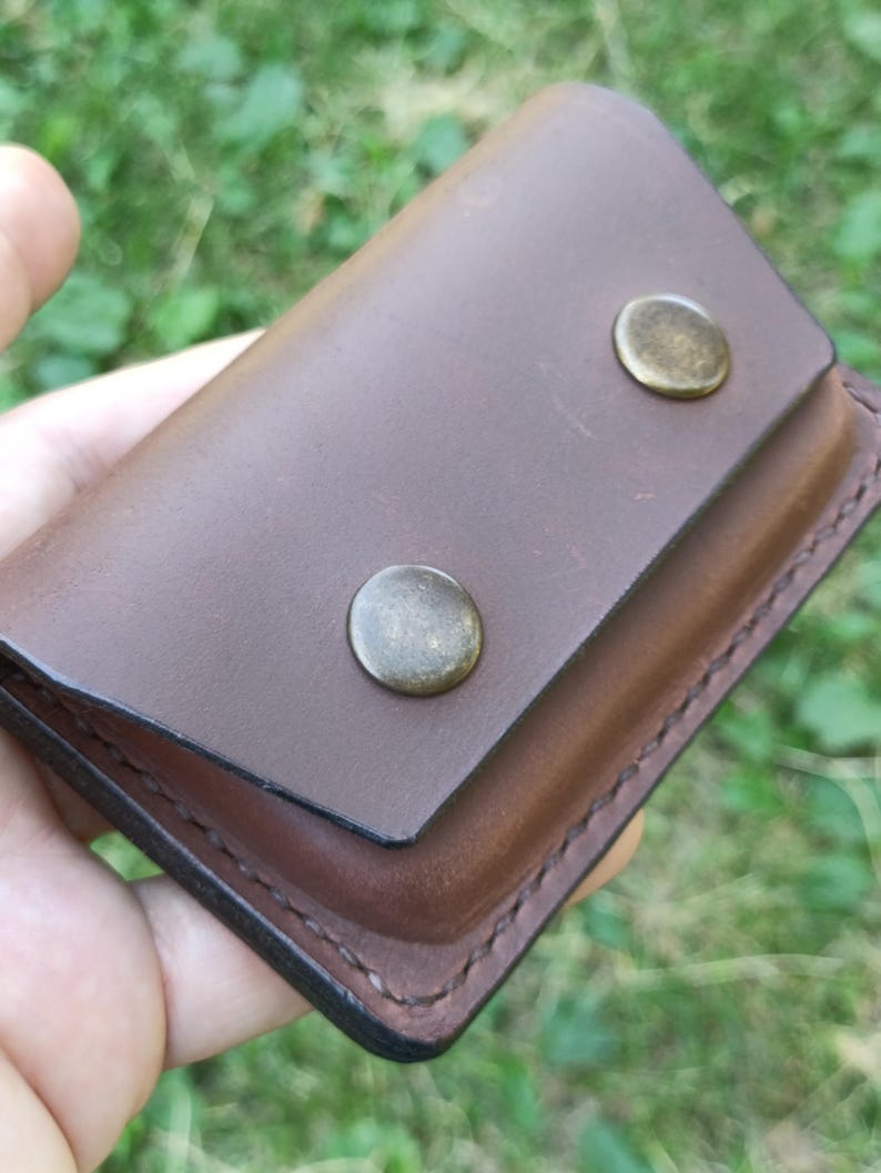 Puede incluir: Un tarjetero rectangular de cuero marr&oacute;n con cierre de solapa asegurado por dos broches met&aacute;licos redondos de color lat&oacute;n. Los bordes del cuero est&aacute;n cosidos con hilo oscuro y la superficie tiene un acabado mate suave con grano visible.