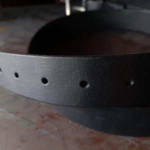 Puede incluir: Un cintur&oacute;n negro con una superficie texturizada y una serie de agujeros circulares espaciados uniformemente a lo largo de la correa. El cintur&oacute;n est&aacute; curvado en un bucle, mostrando el acabado mate oscuro del material y los agujeros.