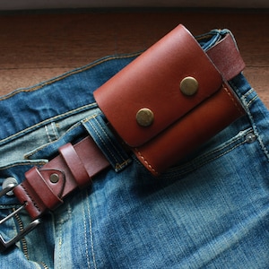 May include: A brown leather wallet with two brass snaps is attached to a brown leather belt. The belt is worn around a pair of blue jeans.