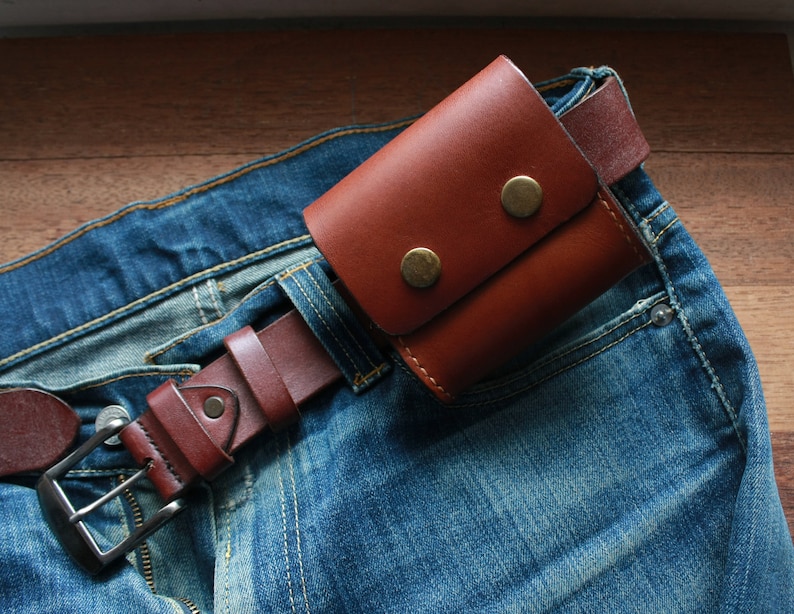 May include: A brown leather wallet with two brass snaps is attached to a brown leather belt. The belt is worn around a pair of blue jeans.