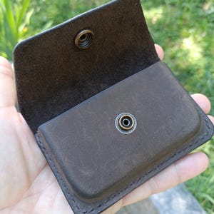 May include: A dark brown leather card holder with a rectangular shape and rounded corners. The accessory features a flap closure secured by a circular antique brass metal snap button fastener centered on the front and the interior flap.