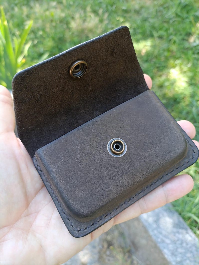 May include: A dark brown leather card holder with a rectangular shape and rounded corners. The accessory features a flap closure secured by a circular antique brass metal snap button fastener centered on the front and the interior flap.