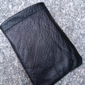 May include: A small, rectangular black leather pouch with a visible pebbled texture. The item features a simple, folded construction with a stitched top edge, presenting a soft, flexible appearance and a deep, matte black finish throughout the surface.