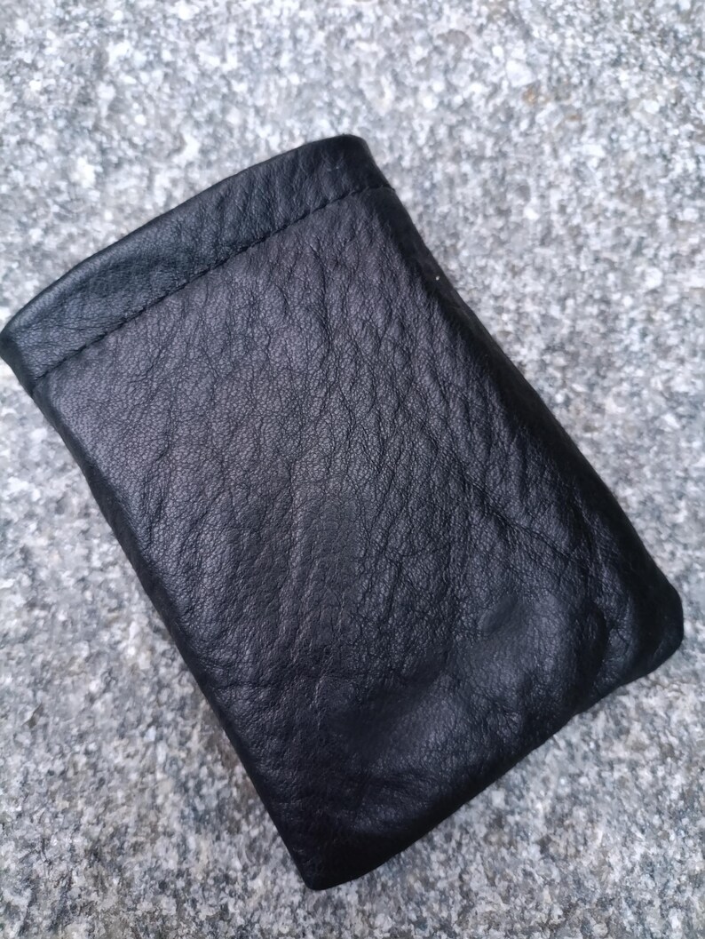 May include: A small, rectangular black leather pouch with a visible pebbled texture. The item features a simple, folded construction with a stitched top edge, presenting a soft, flexible appearance and a deep, matte black finish throughout the surface.