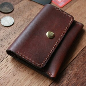 May include: A brown leather wallet with a brass snap closure. The wallet is made of a rich, dark brown leather and features detailed stitching around the edges. The wallet is designed to hold cards and cash. The wallet is approximately 4 inches wide and 3 inches tall.