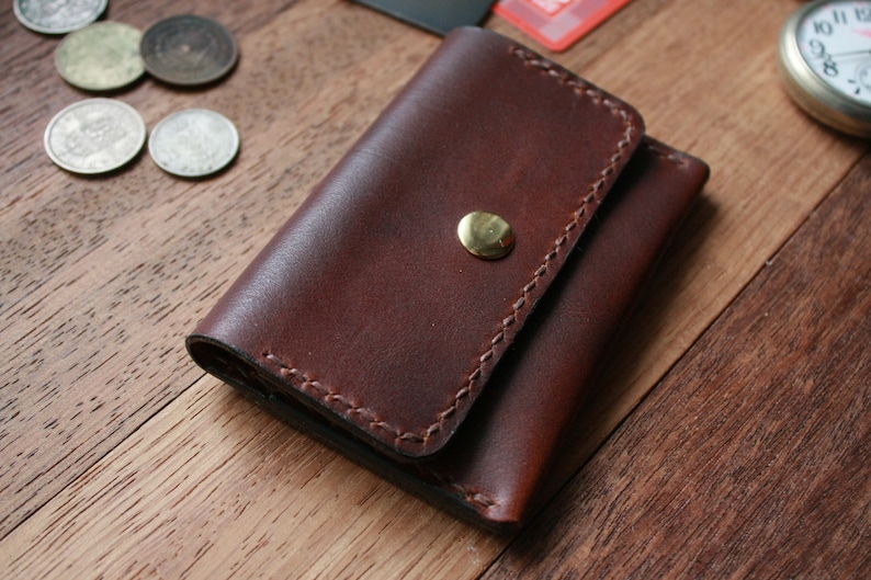 May include: A brown leather wallet with a brass snap closure. The wallet is made of a rich, dark brown leather and features detailed stitching around the edges. The wallet is designed to hold cards and cash. The wallet is approximately 4 inches wide and 3 inches tall.