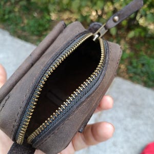 Peut inclure: Une petite pochette en cuir marron fonc&eacute; avec une finition mate et une fermeture &eacute;clair en m&eacute;tal couleur laiton. La pochette pr&eacute;sente une forme rectangulaire aux bords arrondis et une tirette en cuir assortie fix&eacute;e au curseur m&eacute;tallique.