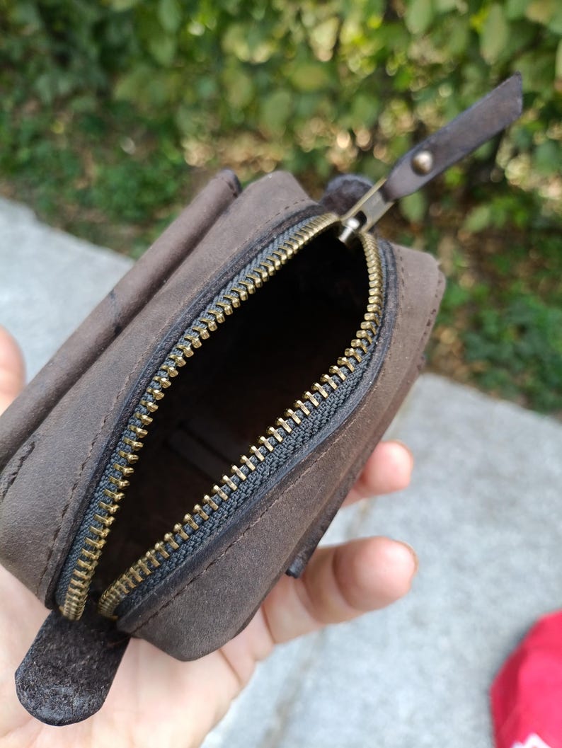 Peut inclure: Une petite pochette en cuir marron fonc&eacute; avec une finition mate et une fermeture &eacute;clair en m&eacute;tal couleur laiton. La pochette pr&eacute;sente une forme rectangulaire aux bords arrondis et une tirette en cuir assortie fix&eacute;e au curseur m&eacute;tallique.