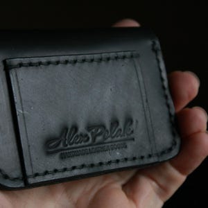 Puede incluir: Cartera de cuero negro con borde cosido y el texto "Alex Polak Handmade Leather Goods" grabado en relieve en la parte delantera.
