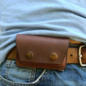 Puede incluir: Una funda rectangular de cuero marr&oacute;n con cierre de solapa asegurado por dos broches de metal color lat&oacute;n. La funda est&aacute; sujeta a un cintur&oacute;n de cuero marr&oacute;n que se lleva sobre unos vaqueros azules y un polo de color azul claro.