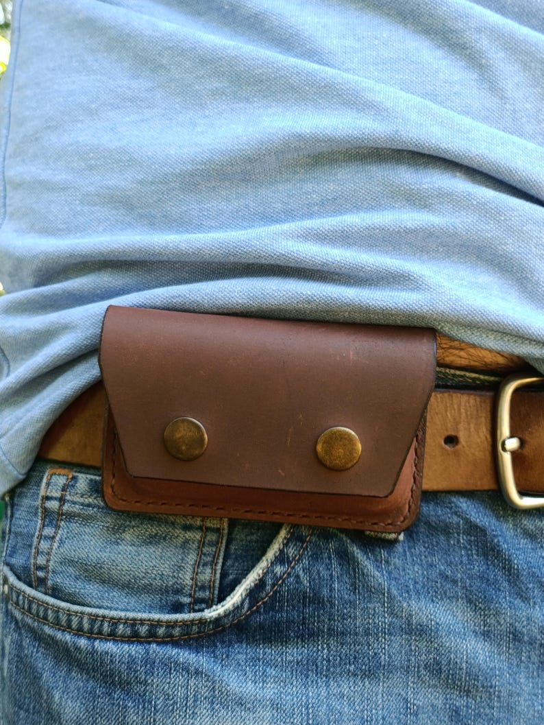 Puede incluir: Una funda rectangular de cuero marr&oacute;n con cierre de solapa asegurado por dos broches de metal color lat&oacute;n. La funda est&aacute; sujeta a un cintur&oacute;n de cuero marr&oacute;n que se lleva sobre unos vaqueros azules y un polo de color azul claro.