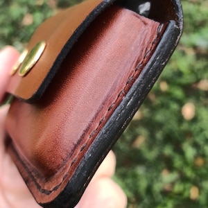 May include: A brown leather pouch featuring a smooth finish, dark burnished edges, and visible black stitching along the side. The pouch is secured with a metallic gold-colored snap button closure on the front flap, showing a rounded rectangular shape.