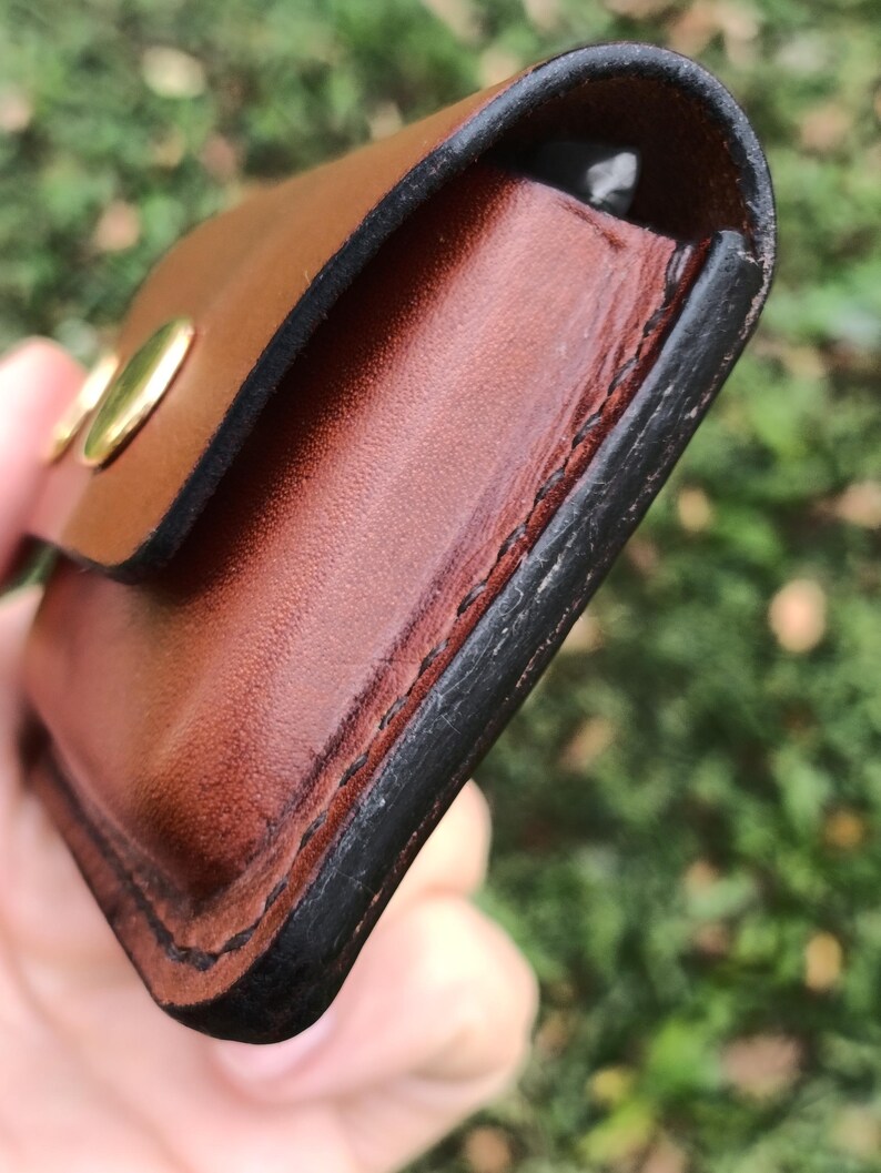 May include: A brown leather pouch featuring a smooth finish, dark burnished edges, and visible black stitching along the side. The pouch is secured with a metallic gold-colored snap button closure on the front flap, showing a rounded rectangular shape.