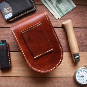 May include: A collection of leather goods, including a brown leather money clip with white stitching, a black wallet, and a small black leather case. A pocket knife and a silver pocket watch are also present.