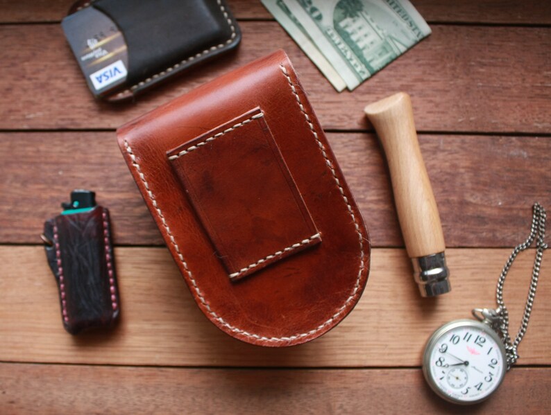 May include: A collection of leather goods, including a brown leather money clip with white stitching, a black wallet, and a small black leather case. A pocket knife and a silver pocket watch are also present.