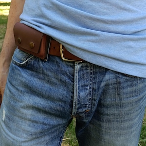 Puede incluir: Un cintur&oacute;n de cuero marr&oacute;n con una bolsa rectangular sujeta en el lateral. La bolsa est&aacute; asegurada con dos botones de presi&oacute;n met&aacute;licos de color lat&oacute;n. El cintur&oacute;n se abrocha con una hebilla met&aacute;lica sencilla y se lleva sobre unos vaqueros azules.