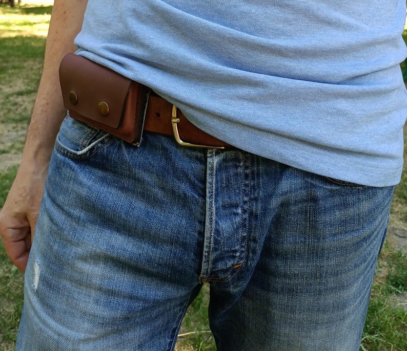 Puede incluir: Un cintur&oacute;n de cuero marr&oacute;n con una bolsa rectangular sujeta en el lateral. La bolsa est&aacute; asegurada con dos botones de presi&oacute;n met&aacute;licos de color lat&oacute;n. El cintur&oacute;n se abrocha con una hebilla met&aacute;lica sencilla y se lleva sobre unos vaqueros azules.