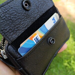 Puede incluir: Una cartera de cuero negro texturizado con cierre de bot&oacute;n a presi&oacute;n. La cartera est&aacute; abierta, revelando una ranura para tarjetas que contiene una tarjeta blanca con elementos gr&aacute;ficos azules, naranjas y amarillos y el texto "KRANK".