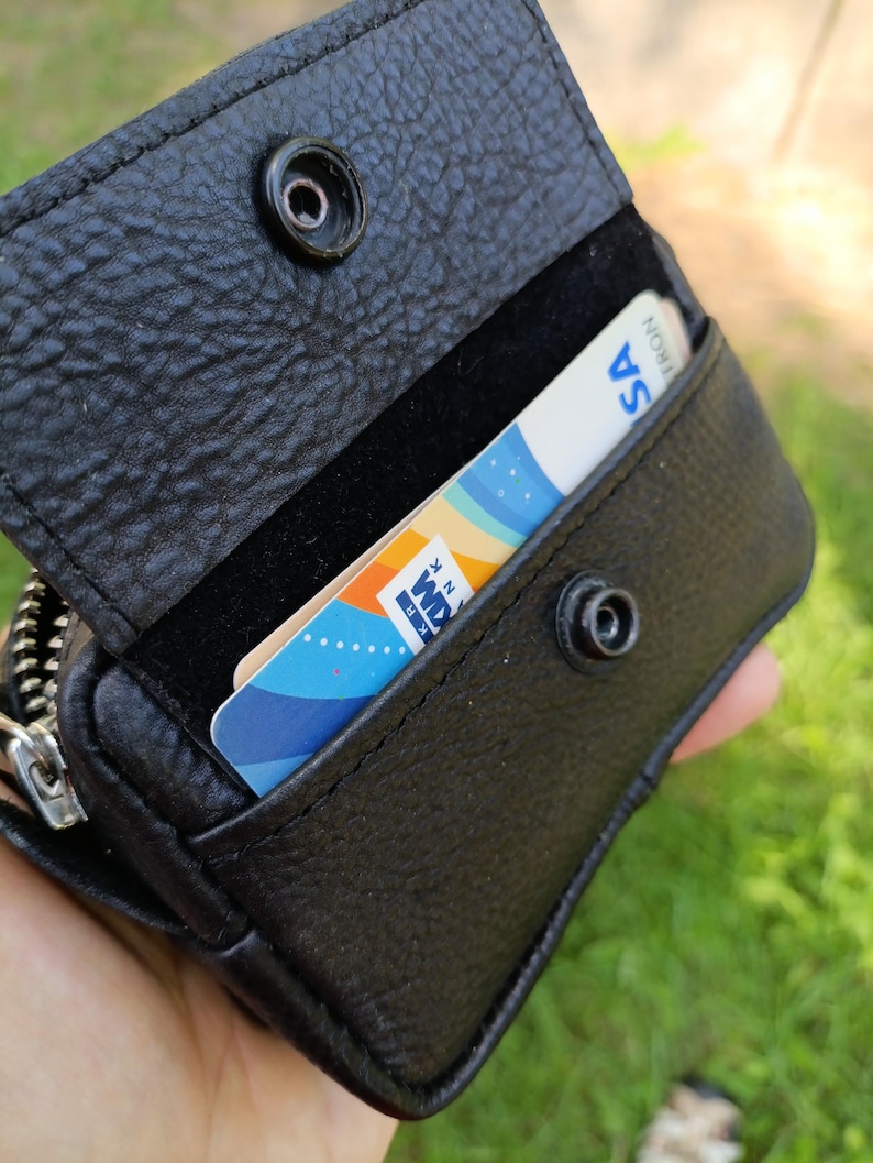 Puede incluir: Una cartera de cuero negro texturizado con cierre de bot&oacute;n a presi&oacute;n. La cartera est&aacute; abierta, revelando una ranura para tarjetas que contiene una tarjeta blanca con elementos gr&aacute;ficos azules, naranjas y amarillos y el texto "KRANK".
