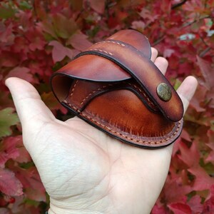 K&ouml;nnte beinhalten: Eine handgefertigte braune Ledertasche mit Klappe und Messingknopfverschluss. Die Tasche wird in einer Hand gehalten, was die detaillierten N&auml;hte und die satte Farbe des Leders zeigt. Der Hintergrund zeigt rotes und gr&uuml;nes Laub.