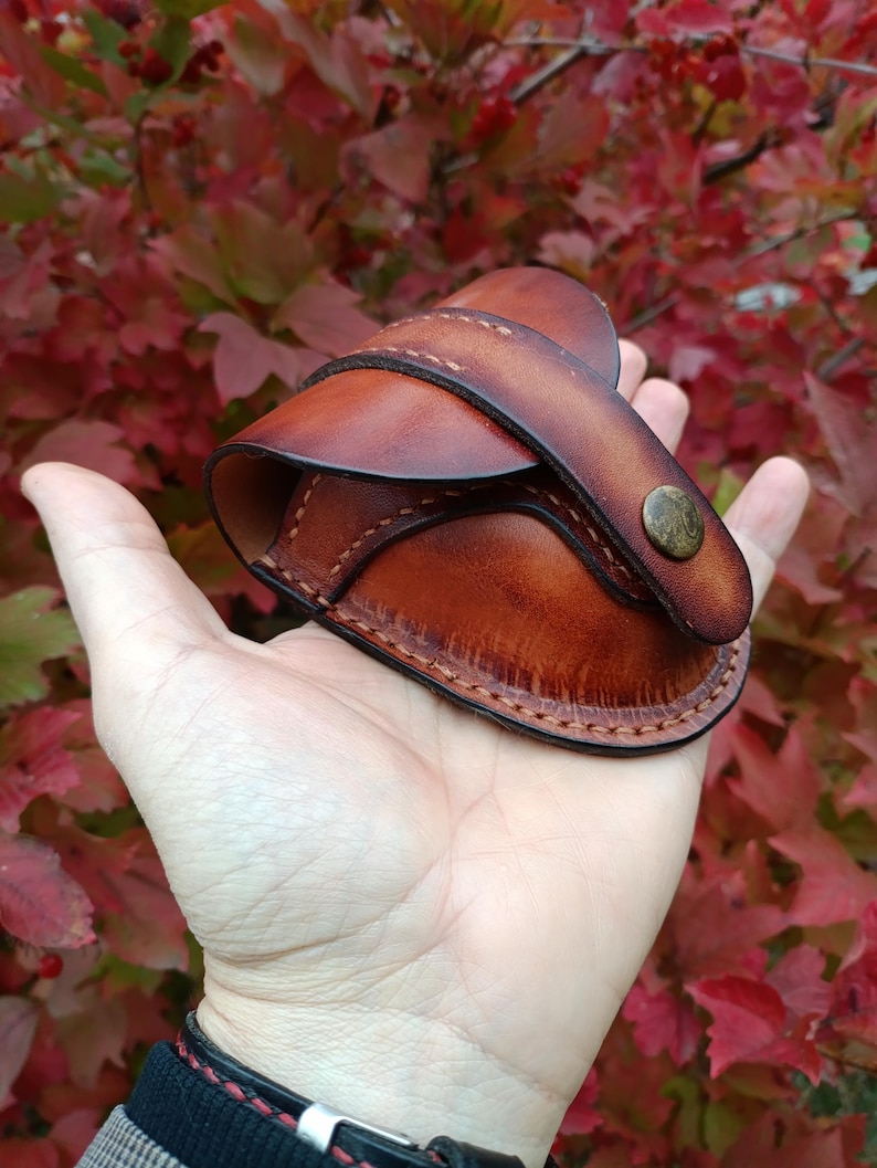 K&ouml;nnte beinhalten: Eine handgefertigte braune Ledertasche mit Klappe und Messingknopfverschluss. Die Tasche wird in einer Hand gehalten, was die detaillierten N&auml;hte und die satte Farbe des Leders zeigt. Der Hintergrund zeigt rotes und gr&uuml;nes Laub.