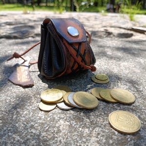 May include: A small, handcrafted leather pouch with a brown and black color scheme, featuring a silver button closure and decorative stitching. Several gold and silver coins are scattered nearby, suggesting a medieval or fantasy theme.