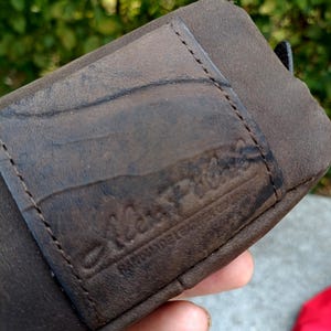 Peut inclure: Un portefeuille en cuir marron fonc&eacute; dot&eacute; d'une poche rectangulaire cousue sur le devant. La surface du cuir pr&eacute;sente une texture grain&eacute;e naturelle et un logo en creux indiquant Alex Piel avec le texte Handmade Leather Goods en dessous.