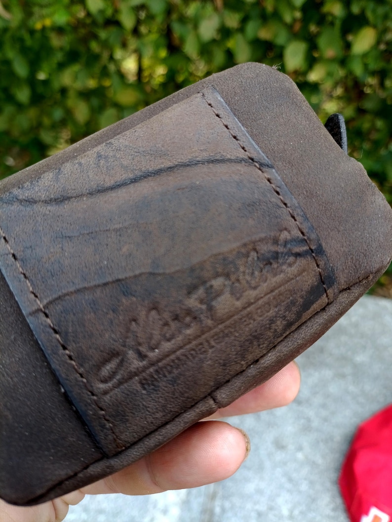 Peut inclure: Un portefeuille en cuir marron fonc&eacute; dot&eacute; d'une poche rectangulaire cousue sur le devant. La surface du cuir pr&eacute;sente une texture grain&eacute;e naturelle et un logo en creux indiquant Alex Piel avec le texte Handmade Leather Goods en dessous.