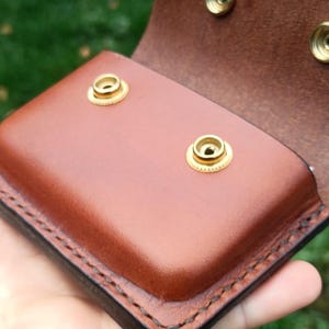 Puede incluir: Una bolsa de cuero marr&oacute;n con cierre de solapa que presenta dos botones de presi&oacute;n de metal dorado en la parte frontal y dos botones a juego en la solapa interior. Los bordes est&aacute;n cosidos con hilo oscuro, mostrando un acabado liso.